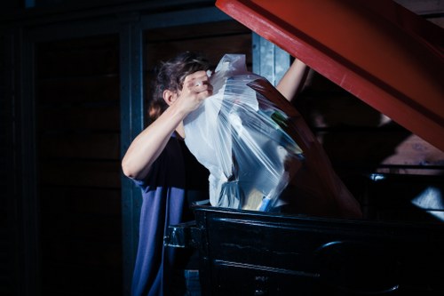 A person wearing a dark T-shirt is placing a clear plastic bag filled with various pieces of waste into a large black dumpster, which is situated outdoors next to a wooden structure. The waste appears to include paper, cardboard, and other household rubbish. Red metal stairs or a platform are visible in the background, partially extending over the scene. The environment suggests a residential or commercial waste disposal area, with the scene lit by natural or artificial light creating shadows on the wooden background. The image is relevant to rubbish clearance services offered by Garden Clearance Barking, highlighting the process of disposing of household and garden waste efficiently.