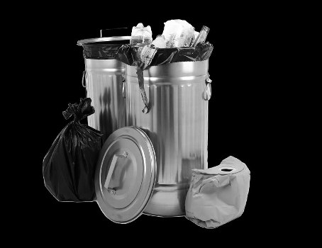 A large, cylindrical metal rubbish bin with a shiny, reflective surface is filled with crumpled paper, plastic bottles, and discarded packaging, indicating general waste. The bin has a closed, domed lid placed next to it, which features a handle for opening. To the left of the bin, there is a tied black plastic waste bag, appearing full and resting on the ground. To the right, a white plastic bag with additional rubbish is visible, sitting on the surface nearby. The scene is set against a plain black background, highlighting the metallic texture of the bin and the variety of waste materials. This setup suggests a typical urban or domestic rubbish collection scenario, relevant to professional waste management services in areas such as Barking. Garden Clearance Barking offers rubbish removal, and this image exemplifies the types of waste items they handle, including paper, plastic, and general household waste, often collected from garden or home clearance in the local area.